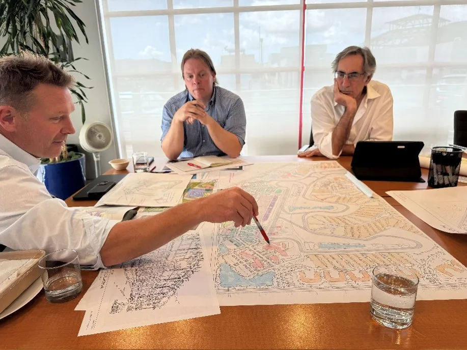 Three colleagues in a working session around a boardroom table, studying a large colour-coded master site plan while one of them marks it with a pen