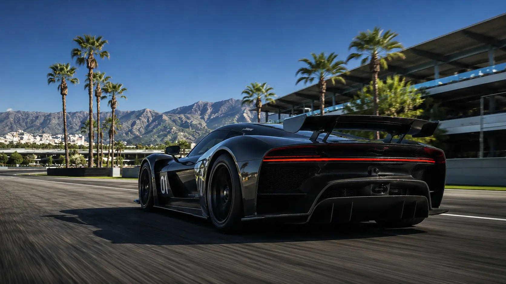 A black competition car at speed on a desert circuit, seen from behind, with palm trees, mountains, and the pit building beyond.